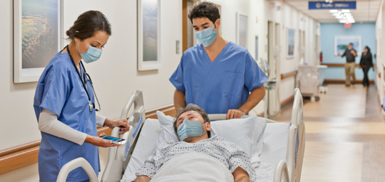 A nurse moving a patient's bed down the hall while another nurse references the patient's information on a Zebra mobile computer